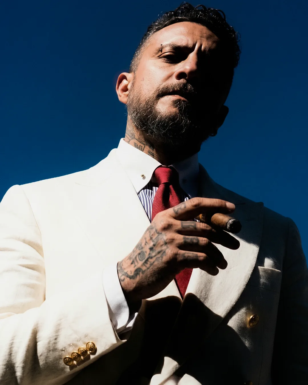 Member gesturing with a cigar, cream suit against a deep blue sky